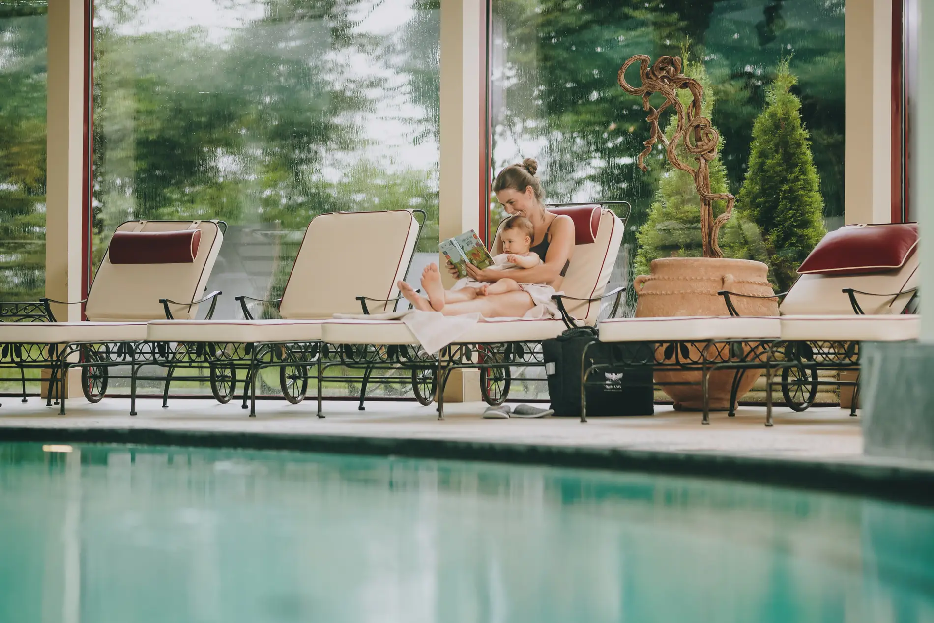 Frau sitzt mit ihrem Baby im Schoß auf einer Liege am Pool und liest ein Buch.